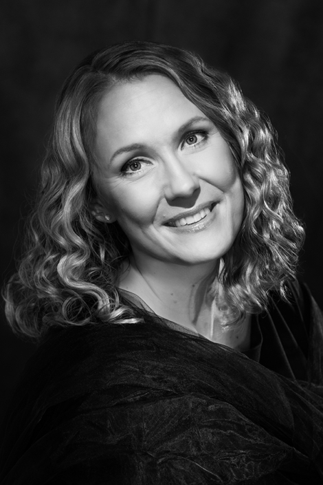 Black and white portrait of a smiling woman with curly hair, wearing a dark textured fabric, captured against a soft background with elegant lighting.