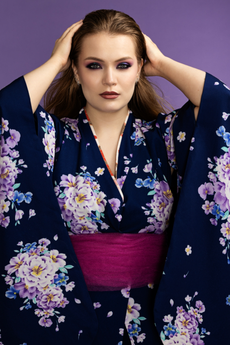 Studiokuvassa nuori nainen katsoo suoraan kameraan kädet hiuksissaan. Hänellä on yllään tumma, kukkakuvioinen kimono, jossa on violetteja ja vaaleita kukkia sekä leveä punertava vyö. Taustana on tasainen violetti studio-tausta, ja meikki korostaa erityisesti silmiä tummilla ja violetin sävyillä.