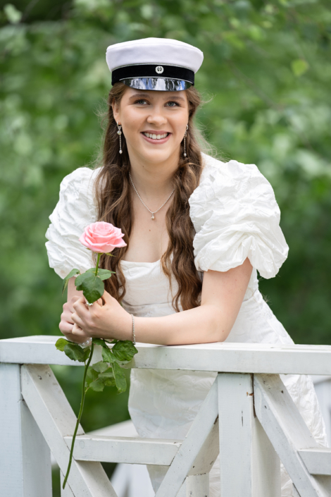 Ylioppilaskuvaus, Hakunilan kartanon puisto Ylioppilas valkoisessa mekossa ja ylioppilaslakissa pinkin ruusun kanssa ulkona. Graduate in a white dress and graduation cap holding a pink rose outdoors.
