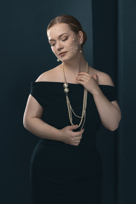Nainen mustassa off-shoulder-mekossa, helminauha ja helmet kasvoilla, tumma sininen tausta. Woman in a black off-shoulder dress with pearl necklace and pearls on her face against a dark blue background.