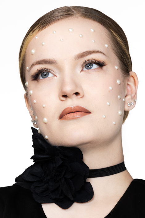 Lähikuva naisesta, helmikoristeet kasvoilla, musta kukkakaulakoru vaaleaa taustaa vasten. Close-up portrait of a woman with pearl embellishments on her face and a black floral choker against a light background.