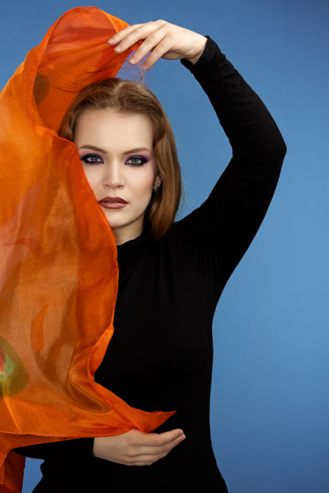 Muotokuva naisesta mustassa yläosassa sinistä taustaa vasten, pitelemässä läpikuultavaa oranssia kangasta kasvojensa edessä; voimakas silmämeikki ja katse suoraan kameraan. Portrait of a woman in a black top against a blue background, holding a translucent orange fabric in front of her face; bold eye makeup and direct gaze at the camera.