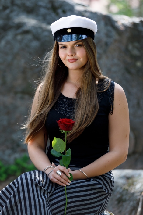 Ylioppilas, kuvaus Espoossa Pitkätukkainen nuori nainen istuu kivellä, pitää kädessään punaista ruusua ja on pukeutunut mustaan toppiin ja raidallisiin housuihin. Päässään hänellä on valkoinen ylioppilaslakki.